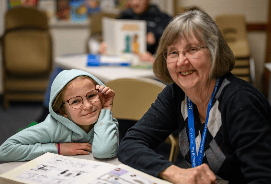 Young student wearing glasses sitting with an adult mentor while completing a worksheet at a table