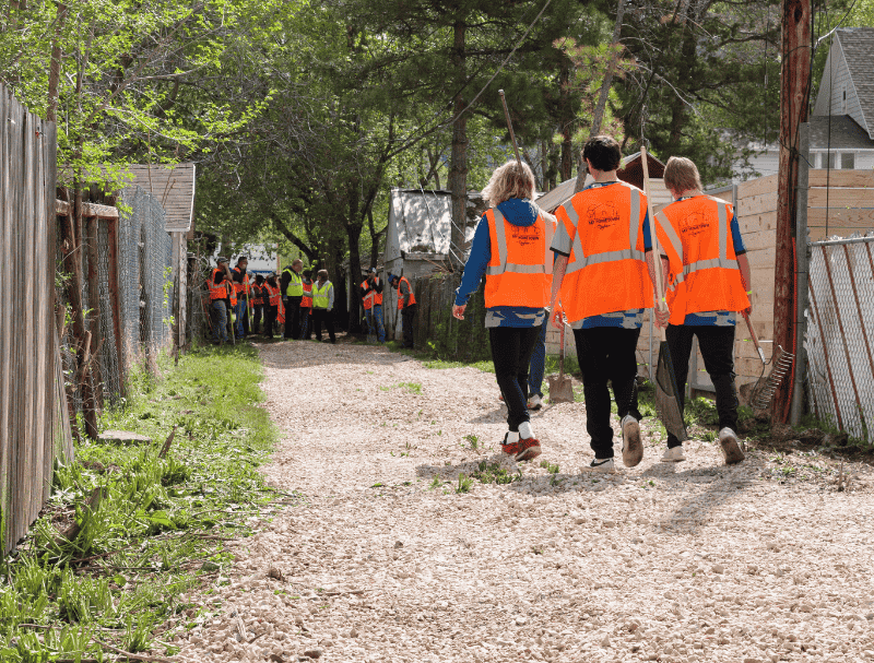 Volunteers wearing bright orange safety vests walk down a gravel alley toward a larger group gathered for a neighborhood cleanup project in Utah.