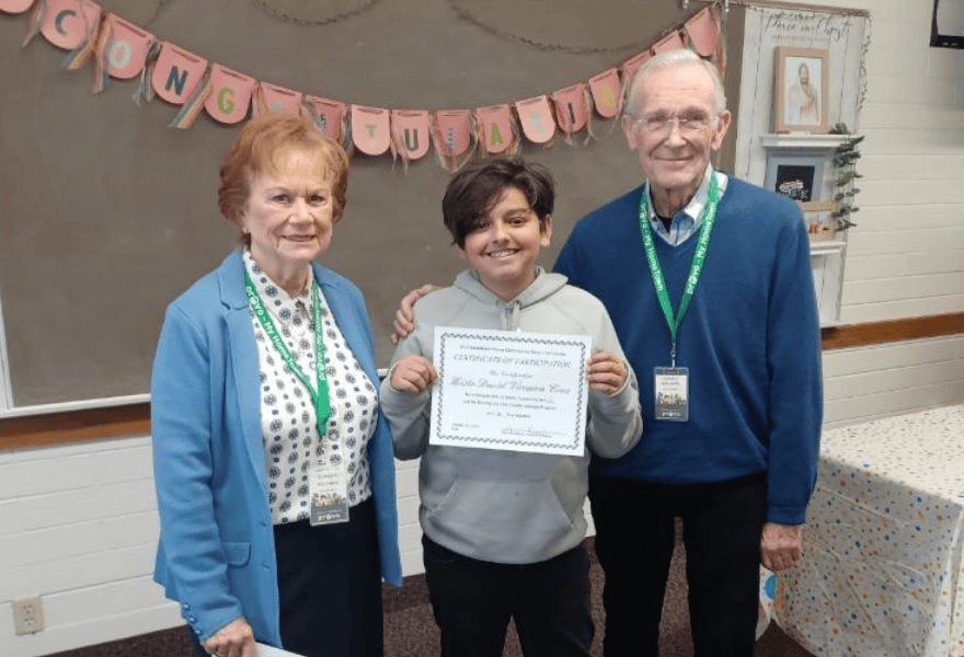 Student smiling and holding an award certificate while standing with two adults in a classroom