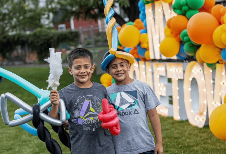 Two boys smiling at a community festival holding cotton candy and balloon creations with colorful decorations behind them