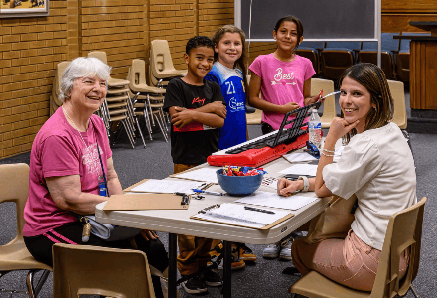 Group of kids standing by a keyboard while two volunteers lead a music lesson and activity