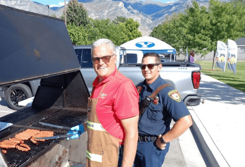 Two men smiling while grilling at a community gathering with tents and mountains behind them
