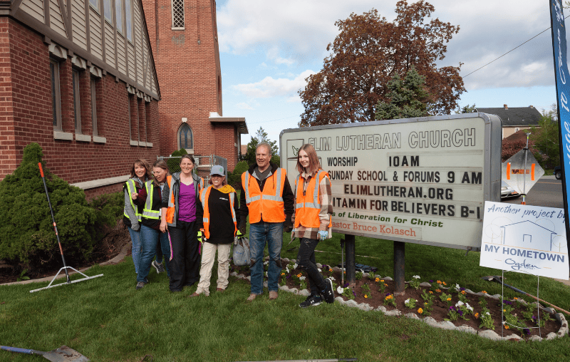 Volunteers in orange safety vests stand together on a church lawn after completing landscaping work during a Utah neighborhood revitalization project.