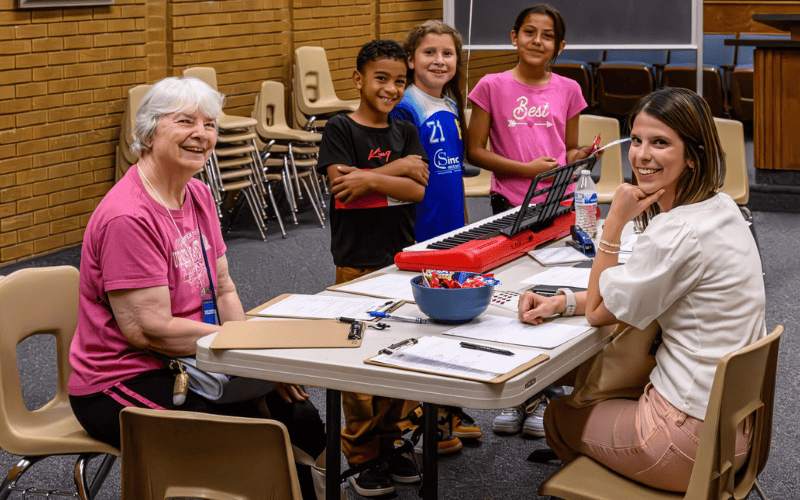 Group of kids standing by a keyboard while two volunteers lead a music lesson and activity