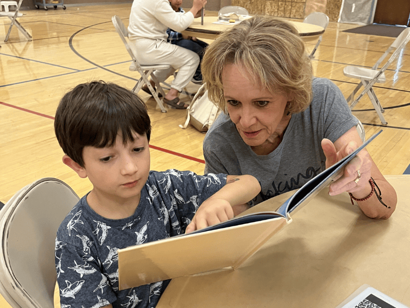 Adult mentor guiding a child through reading during a literacy activity in a gymnasium setting