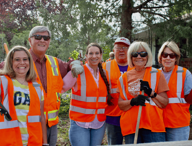A group of smiling volunteers in orange safety vests stand together outdoors with gardening tools after completing a neighborhood service project.