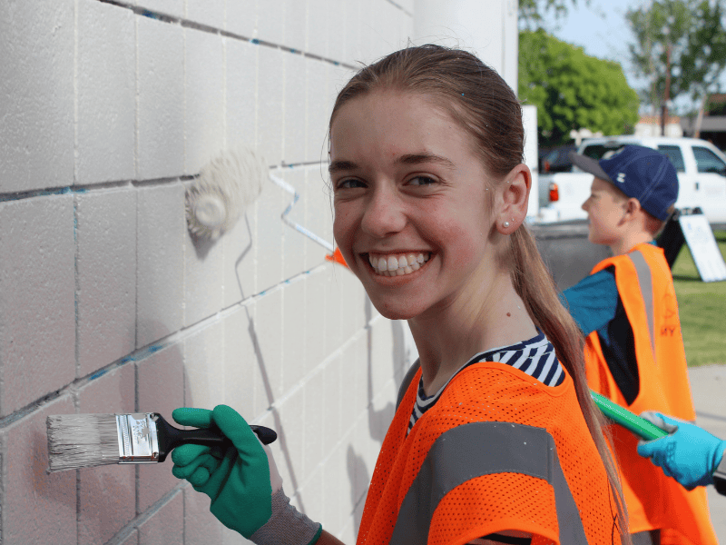 Volunteer in a safety vest painting a building wall alongside others during a community project