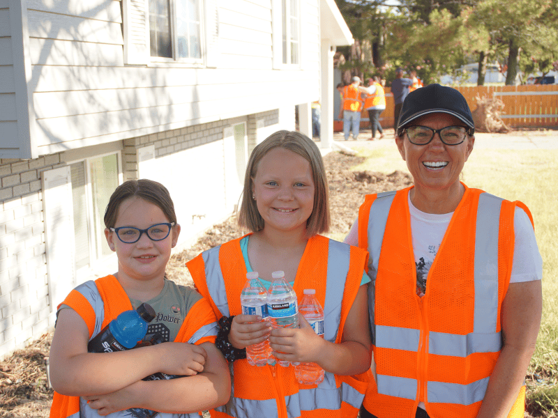 Volunteer and two children wearing safety vests holding water bottles at an outdoor service activity