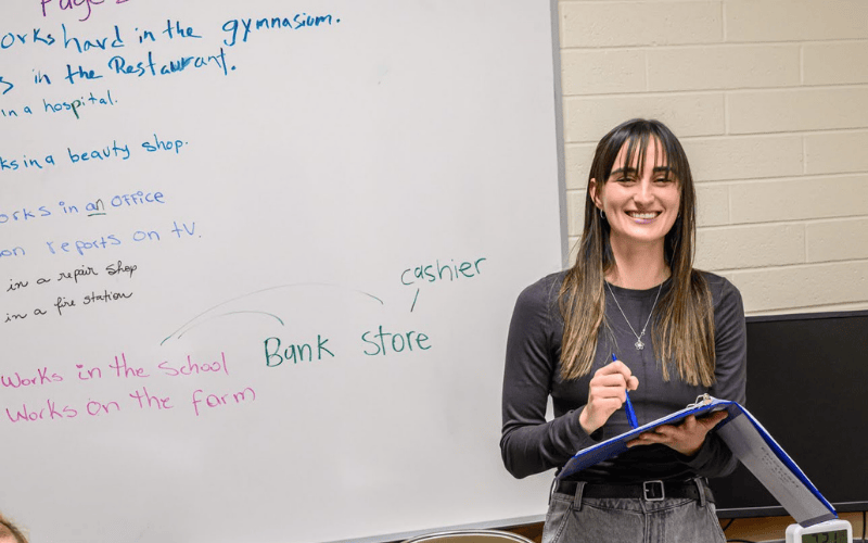 Teacher holding a clipboard and smiling beside a whiteboard with lesson notes about careers and job locations