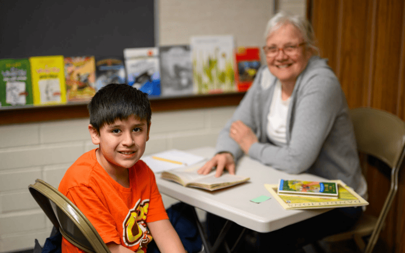 Child sitting at a table with a mentor while reading books together in a classroom setting
