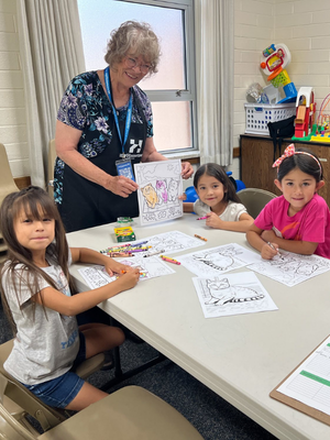 Teacher assisting children as they color worksheets together at a table in a classroom setting
